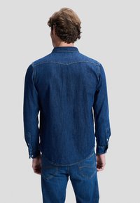 Vyras su bangomis plaukuose, vilkintis ilgomis rankovėmis mėlyną džinsinę marškinių ir atitinkančių mėlynų džinsinių kelnių komplektą, matomas iš nugaros.