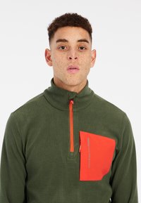 Groene pullover met een gestructureerde afwerking, met een oranje halve rits en een contrasterende oranje borstzak met tekst langs de rand.