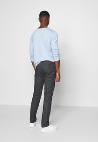 Mężczyzna stojący tyłem, ubrany w jasnoniebieską koszulę z długim rękawem, ciemnoszare spodnie i białe sneakersy, na jednolitym białym tle.