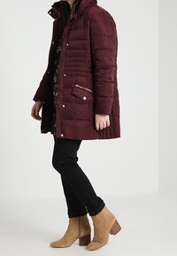 Cappotto imbottito bordeaux con tasca con cerniera e chiusure a pressione, abbinato a pantaloni neri aderenti e stivaletti beige con tacco a blocco.