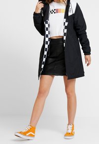 Person som bär en svart lång jacka med rutigt foder, en vit kort ärmad grafisk t-shirt, en svart läder minikjol och orangea hög-top sneakers.