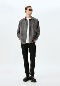 Grå ternet skjorte med en frontlomme, båret over en hvid t-shirt, kombineret med sorte jeans og sorte kraftige støvler, accessoriseret med solbriller.