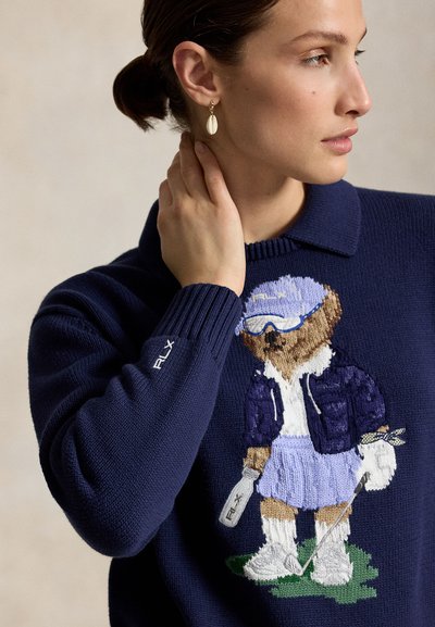 Femme portant un pull bleu marine avec un ours de golf brodé, touchant son cou, et une boucle d’oreille dorée en forme de coquillage.