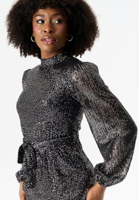 Black sequined dress with high collar, long puffy sleeves, and a tied waist. Shiny textures create a shimmering effect.