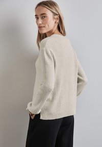 Helle beige, gerippte Sweater mit langen Ärmeln, rundem Ausschnitt und seitlichen Schlitzen. Getragen mit dunklen Hosen aus einem strukturierten, weichen Stoff.