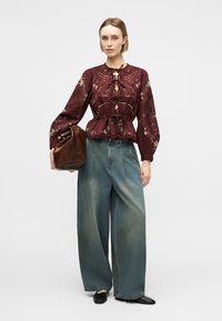 Vrouw die een bordeauxrode blouse met bloemenborduursel en strikjes draagt, wijd uitlopende vervaagde jeans, zwarte schoenen en een grote bruine leren tas vasthoudt.