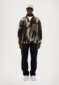 Giacca in peluche con motivo camouflage nei colori verde, beige e nero, con una tasca con zip, abbinata a pantaloni scuri e sneakers bianche.