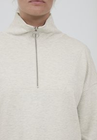 Lysegrå fleecepullover med høj krave, der har en halv lynlås og struktureret overflade. Sat mod en neutral baggrund.