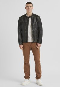 Schwarze Lederjacke mit gesteppten Schulterakzenten, kombiniert mit einem beigen Pullover, hellbraunen Hosen und weißen Sneakers. Einfache Reißverschlussdetails.