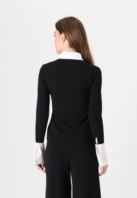 Svart langermet topp med hvit krage og satengensker, med en tettsittende design og subtile splitt ved ermene. Glatt tekstur.