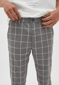 Mani che regolano il punto vita di pantaloni a quadri grigi indossati con una maglietta bianca con la scritta "SONI MUSICALI" sul davanti.