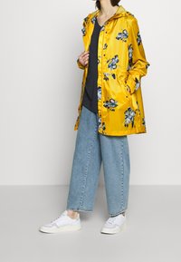 Chubasquero amarillo con estampado floral, que cuenta con capucha, cierres a presión y cintura elástica, combinado con vaqueros azules holgados y zapatillas blancas.