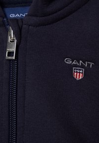 Sudadera con cremallera en color azul marino, hecha de tejido suave, con una cremallera plateada y el logotipo de GANT bordado con un detalle de escudo en rojo y azul.