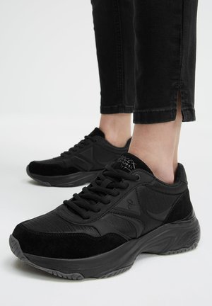 Black chunky sneakers with laces worn with black cropped pants featuring a small slit at the ankle against a white background.