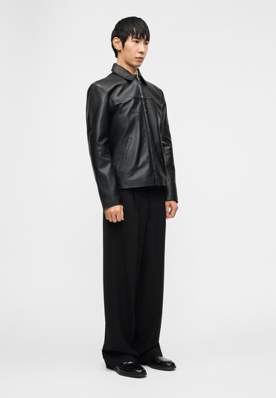 Άνδρας που φοράει μαύρο δερμάτινο μπουφάν, χαλαρό μαύρο παντελόνι και μαύρα παπούτσια, στέκεται μπροστά σε ένα απλό ανοιχτό γκρι φόντο.