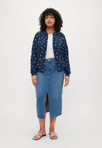 Veste bombardier navy avec imprimé floral, assortie d'une jupe en denim longueur genou avec une fente à l'avant. Le mannequin porte un haut blanc et des sandales.