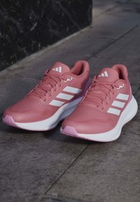 Paire de chaussures de sport roses avec semelles blanches et trois bandes blanches sur les côtés, placées sur un pavé en pierre grise.