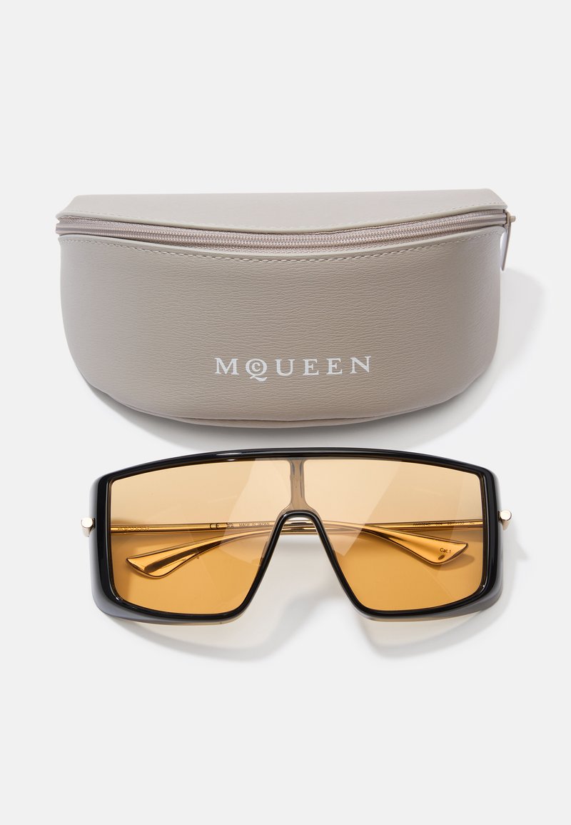 Schwarze übergroße Sonnenbrille mit bernsteinfarbenen Gläsern, rechteckiger Form und goldenen Akzenten. Mit einem grauen Reißverschluss-Etui zur sicheren Aufbewahrung.