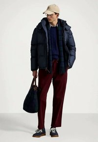 Άνδρας με σακάκι ναυτικού χρώματος, μπλε πουλόβερ, κόκκινο παντελόνι, μπεζ καπέλο, γυαλιά, κρατώντας ναυτικό τσάντα και στέκεται σε λευκό φόντο.