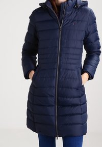 Navy blue puffer coat with a zip front, long sleeves, a hood, and horizontal quilting. Features side pockets and a small logo on the chest.