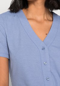 Camicia in tessuto a coste blu con scollatura a V, maniche corte e quattro bottoni sul davanti. La texture è morbida e leggermente strutturata.