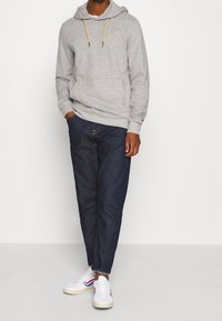 Sudadera con capucha gris con cordones, bolsillo delantero, combinada con vaqueros azul oscuro y zapatillas blancas con detalles en rojo y azul.