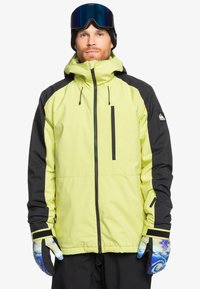 Ljusgul vattentät jacka med svarta accenter, medaljongficka, justerbar huva och en modell som bär färgglada mönstrade handskar.