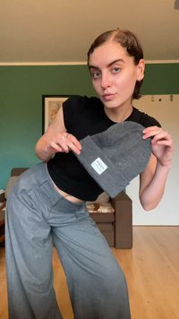 Gorro gris con una etiqueta, sostenido en la mano. Top corto negro y pantalones de pierna ancha grises. Tejido suave, diseño simple y colores neutros.