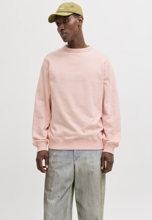 Mees, kes kannab heleroosat ümar-kaelusega pusa, pleekinud halli teksapükse, oliivrohelise mütsi ja väikese rõngakujulise kõrvarõnga, seisab tavalise tausta ees.