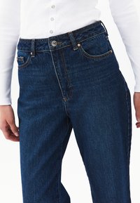 Vaqueros de mezclilla oscura con cintura alta y cinco bolsillos. Presentan costuras en contraste y un cierre de botón metálico. Tejido texturizado con un ligero brillo.