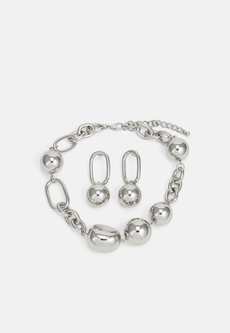 Bracelet ton argent avec de gros maillons de chaîne et des perles rondes, associé à des boucles d'oreilles pendantes à maillons ovales assorties sur fond blanc.
