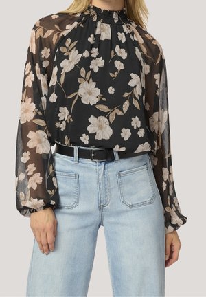 Blouse noire transparente à motif floral, manches longues bouffantes et col haut froncé, rentrée dans un jean bleu clair avec une ceinture noire.