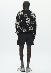 Crni pleteni džemper s kremastim cvjetnim uzorkom, uparen s crnim kratkim hlačama, sivim čarapama i svijetlosivim tenisicama.