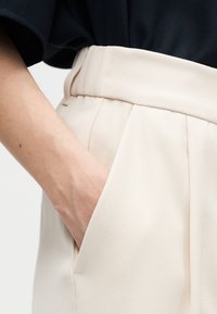 Χέρι ατόμου στην πλαϊνή τσέπη ψηλόμεσου, ανοιχτό μπεζ παντελονιού, σε συνδυασμό με σκουρόχρωμο τοπ φουσκωμένο μέσα.