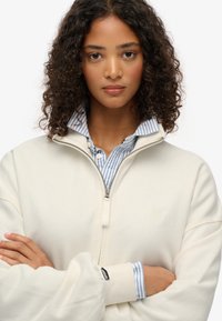 Felpa cremisi con zip e polsini a costine, indossata sopra una camicia a strisce blu e bianche. Presenta un piccolo logo sul petto e una texture liscia.