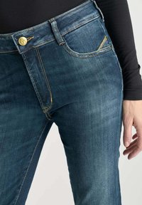 Persona viste unos pantalones de mezclilla azul oscuro y una camiseta negra de manga larga, con la mano descansando al lado del muslo.