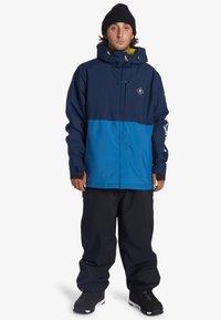 Blå och marinblå jacka med huva, prydd med en diamantlogotyp, elastiska ärmslut och dragkedja framtill. Kombinerad med lösa svarta byxor och stövlar.