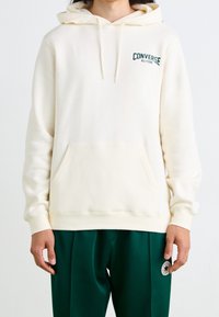 Sweat à capuche couleur crème avec une poche avant, une capuche à cordon de serrage, et l'inscription "Converse All-Star" brodée en vert foncé sur la poitrine.