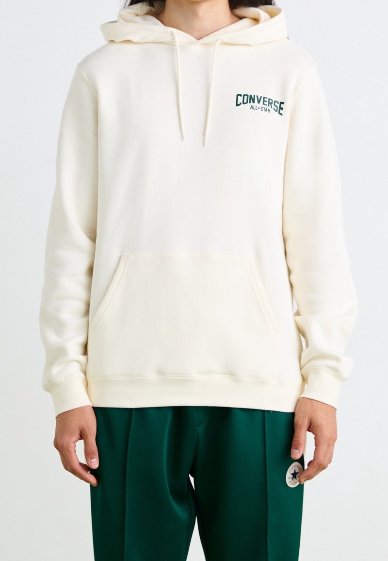 Sweat à capuche couleur crème avec une poche avant, une capuche à cordon de serrage, et l'inscription "Converse All-Star" brodée en vert foncé sur la poitrine.