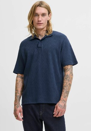 Mörkblå polo-tröja med en texturerad geometrisk mönster, korta ärmar och en krage med två knappar. Bärs med mörka denimbyxor.
