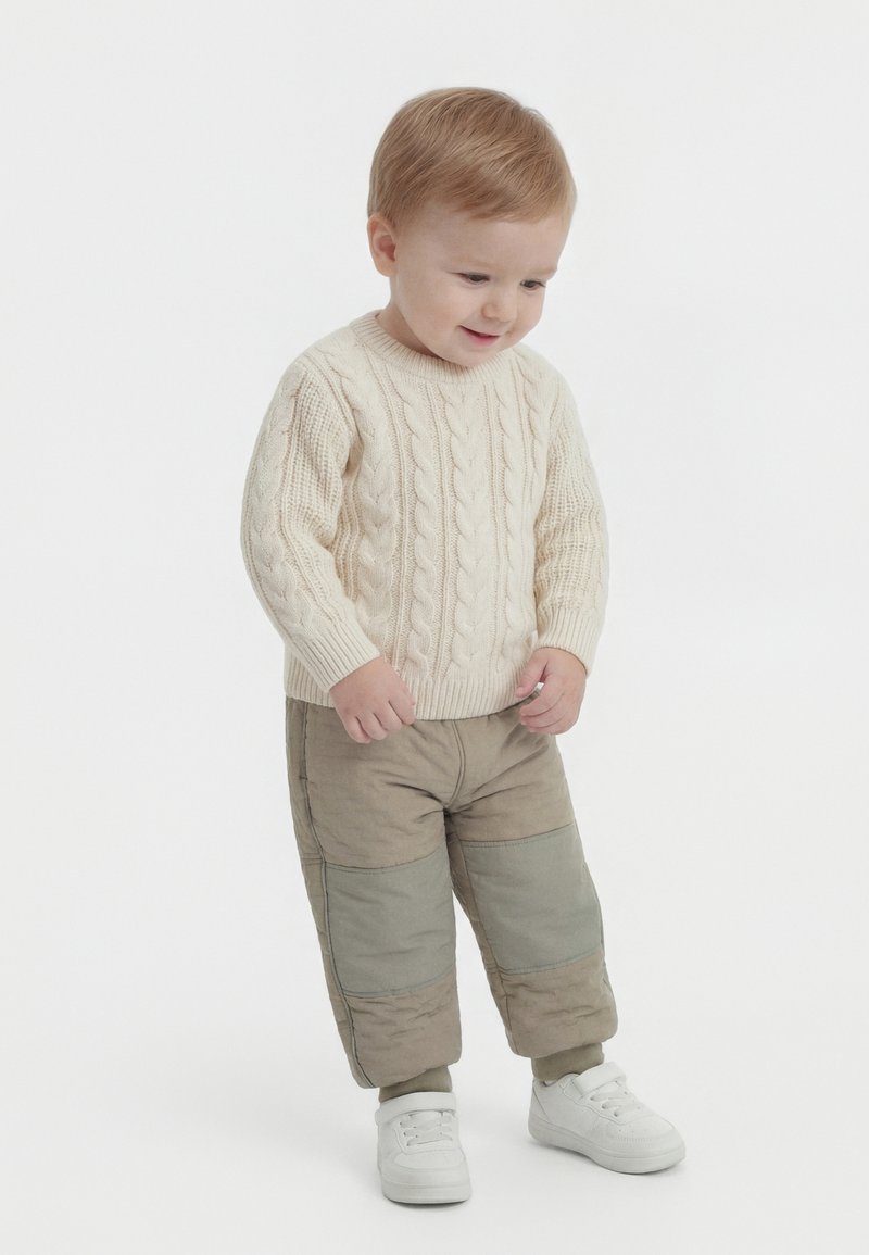 Νεαρό παιδί με μπεζ πουλόβερ πλεκτό με πλεξούδες, παντελόνι πατςγουορκ και λευκά αθλητικά παπούτσια, στέκεται και κοιτάζει κάτω με ελαφρύ χαμόγελο.