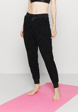 Femme debout pieds nus sur un tapis de yoga rose portant un pantalon jogger à imprimé animalier noir et un soutien-gorge de sport noir.