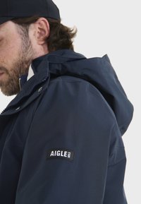 Giacca impermeabile blu navy con cappuccio, caratterizzata da una cerniera frontale, bottoni a pressione e un'etichetta nera con testo bianco sulla manica sinistra.
