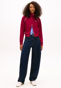 Μπορντό μπουφάν με φερμουάρ και λευκές λεπτομέρειες, με μικρό λογότυπο, σε συνδυασμό με φαρδιά σκούρα τζιν και λευκά αθλητικά παπούτσια.