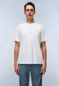 White cotton t-shirt with a crew neck, short sleeves, and a small logo detail on the chest. Paired with light blue trousers.