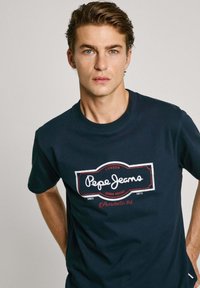 Tamno plava pamučna majica s velikim bijelim i crvenim logotipom "Pepe Jeans" sprijeda, s okruglim izrezom i kratkim rukavima.