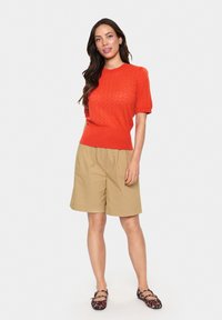 Orange kortärmad stickad topp med ett texturerat mönster, i kombination med khaki veckade shorts. Svart och beige mönstrade ballerina skor. Neutral bakgrund.