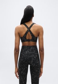 Γυναίκα που φαίνεται από πίσω, φορώντας μαύρο και γκρι κολάν με σχέδιο και ψηλή μέση, καθώς και ασορτί αθλητικό σουτιέν με σταυρωτούς ιμάντες και κομμένη πίσω πλευρά.