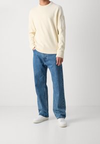 Světlý béžový pletený svetr s kulatým výstřihem a kapsou na rukávu na zip, v kombinaci s volnými modrými džínami a bílými teniskami.
