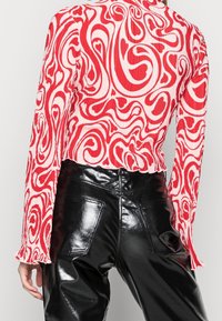 Röd och vit randig långärmad topp med ett virvlande mönster, i kombination med hög midja svarta glänsande byxor. En enfärgad bakgrund.